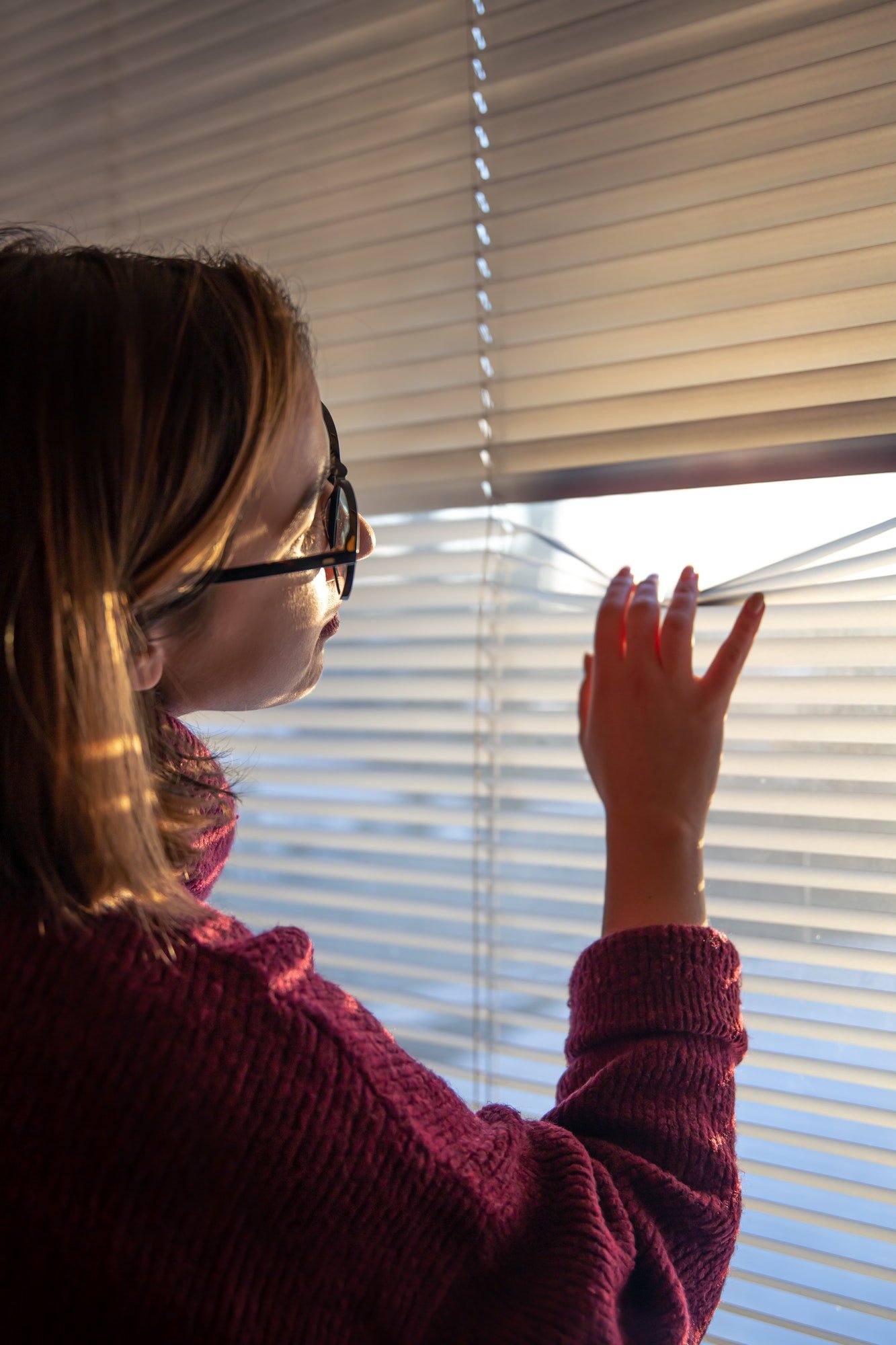 A woman looks through the blinds at the early morning sunlight.